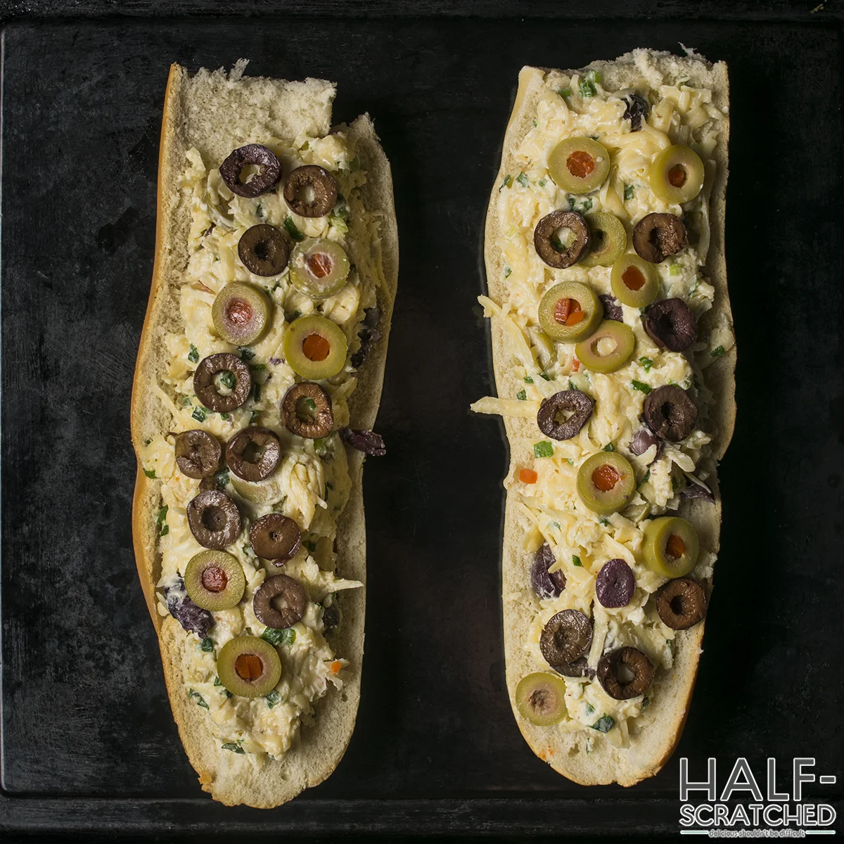 Two halves of olive bread with cheese and olives on a baking sheet